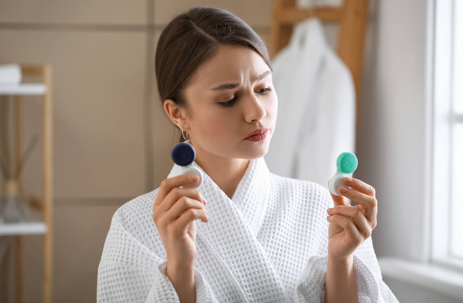 Young woman in her robe holding up two contact lens containers and trying to decide.