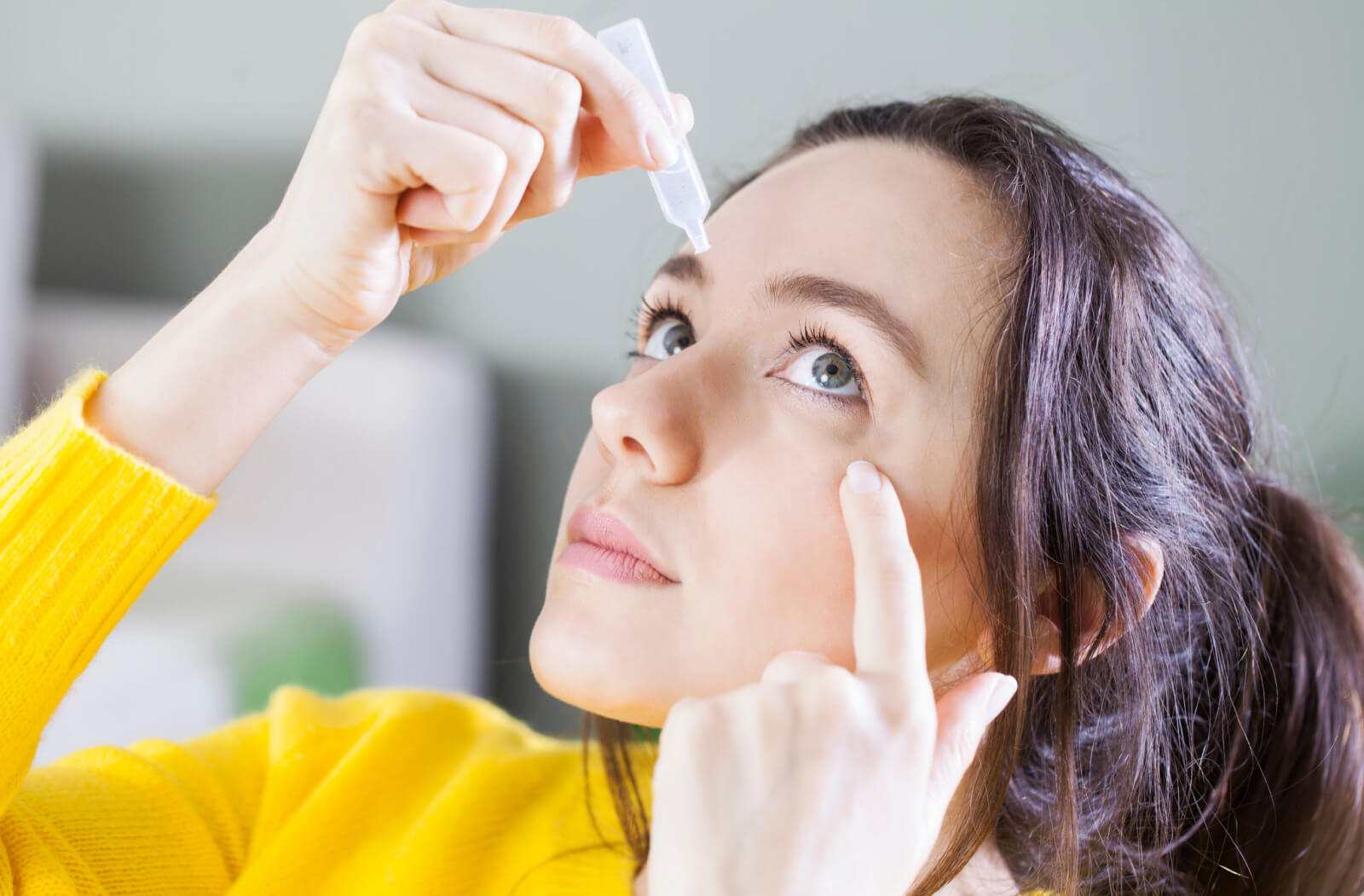 A lady in a yellow shirt holding a small plastic dropper near her eye and dropping a solution lubricating her eye.